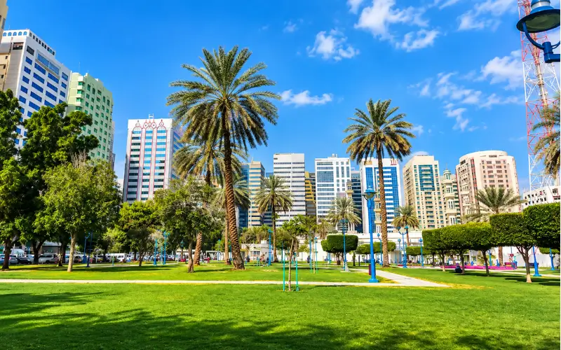 Electra Park with green lawns, palm trees, blue lamp posts, and a modern city skyline.