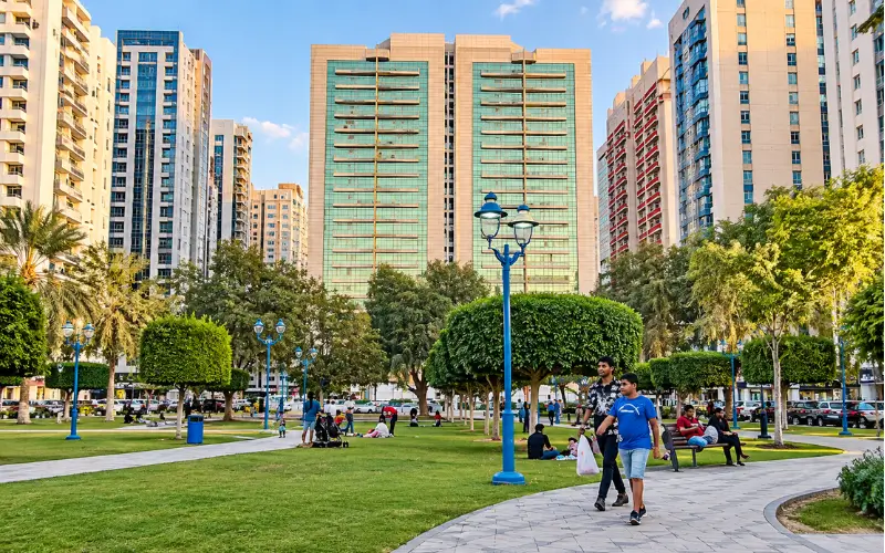 Electra Park with landscaped green lawns, palm trees, walking paths, blue lamp posts, and a modern Abu Dhabi skyline.